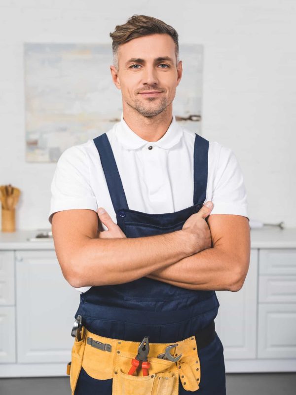 adult repairman in tool belt standing with crossed arms at kitchen and looking at camera